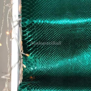 Op de afbeelding: Een levendig smaragdgroen materiaal met een gestructureerd slangenleerpatroon en een glanzende afwerking. Een wit frame begrenst de linkerkant, versierd met een lichtsnoer van kleine, warme lampjes. De tekst AcalopatiBali is zichtbaar.