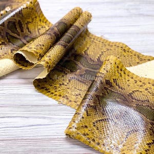 May include: A long, flexible strip of snakeskin leather featuring a repeating diamond pattern in shades of mustard yellow, olive green, and dark brown. The material has a glossy, textured surface with visible scale details and a soft, light-coloured underside.