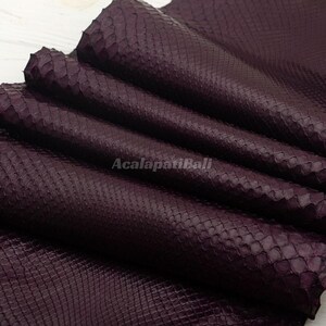 Peut inclure: Une peau de cuir de serpent violet foncé présentant un motif d'écailles répétitif distinct sur toute la surface. Le matériau est plié en couches, mettant en valeur la texture et le texte AcalapatiBali imprimé au centre du matériau.