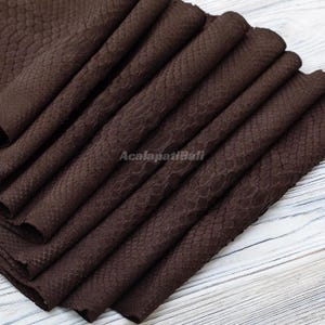 May include: A dark brown leather sheet with a textured snakeskin pattern featuring a repeating scale design. The material is folded in multiple layers, creating deep pleats across the surface. The text AcalapatiBali is printed in the center.