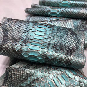 Può includere: Una pila di pezzi rettangolari in pelle di serpente turchese e nera. Ogni pezzo presenta un distinto motivo a scaglie sovrapposte con una finitura lucida, mostrando una sfumatura di vivaci toni blu-verdi che sfumano verso bordi color carbone.