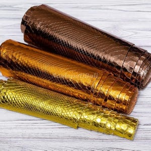 May include: Three rolls of metallic faux leather material featuring a textured snakeskin pattern. The top roll is a deep bronze, the middle roll is a warm copper, and the bottom roll is a bright, reflective gold finish.