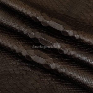 Peut inclure: Un cuir de peau de serpent brun foncé présentant un motif d'écailles répétitif distinct sur toute la surface. La texture est superposée avec des plis, et le texte AcalapatiBali est imprimé dans une police claire et semi-transparente au centre.