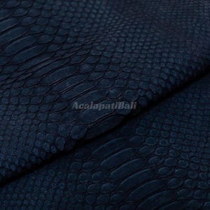 Op de afbeelding: Een donker marineblauw materiaal met een gestructureerd slangenleerpatroon met overlappende schubben. Het oppervlak toont een herhalend organisch schubbenpatroon over het hele stuk. De tekst AcalapatiBali staat in het midden afgedrukt.