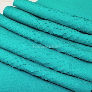 Peut inclure: Une feuille de cuir de serpent de couleur turquoise présentant un motif d'écailles en relief sur toute la surface. Le matériau est plié en plusieurs couches et le texte AcalapatiBali est imprimé en couleur claire au centre.