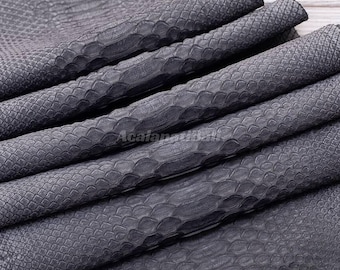 May include: A dark grey material with a textured snakeskin pattern featuring overlapping scales. The surface has a matte finish and is folded into multiple layers, creating a series of horizontal ridges across the entire piece of material.