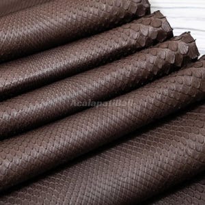 Peut inclure: Un matériau texturé brun foncé avec un motif d'écailles répétitif plié en plusieurs couches. La surface présente un design gaufré à petite échelle cohérent sur tout le tissu. Le texte AcalapatiBali est visible au centre.