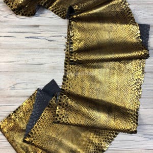 Op de afbeelding: Een stuk metallic goudkleurig leer met een gestructureerd slangenleerpatroon. Het materiaal heeft een herhalend schubbenontwerp met een reflecterende afwerking en een donkergrijze onderkant zichtbaar langs de gevouwen randen.