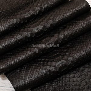 Peut inclure: Un matériau texturé brun foncé présentant un motif répétitif de peau de serpent avec une finition brillante. Le matériau est plié en couches, créant des ombres et des reflets profonds sur la surface. Le texte AcalapatiBali est visible au centre.