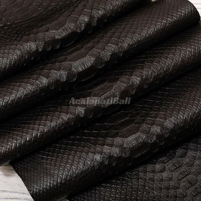 May include: A dark brown, textured material featuring a distinct snakeskin pattern with overlapping scales. The surface has a matte finish and is folded in layers, displaying the intricate, repeating scale design throughout the entire piece.