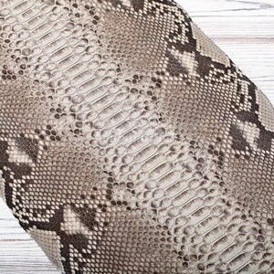 May include: A textured material featuring a realistic snakeskin pattern in shades of beige, cream, and dark brown. The surface displays a repeating scale design with a central row of larger, elongated scales flanked by smaller, rounded scales.