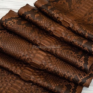 Peut inclure: Une pile de feuilles de cuir de serpent pliées de couleur marron avec un motif d'écailles en losange marron foncé et beige. La surface présente un aspect reptilien texturé avec une finition brillante. Le texte AcalapatiBall est visible au centre.