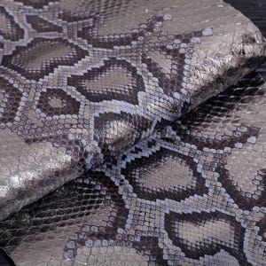 Peut inclure: Une pièce de tissu pliée présentant un motif de peau de serpent gris foncé et argent métallisé. La surface affiche un design texturé réaliste avec des écailles et un motif répétitif en forme de losange sur toute la zone visible.