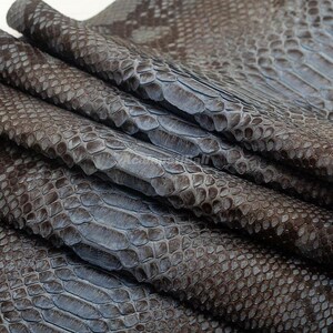 Peut inclure: Une peau de serpent en cuir brun foncé présentant un motif d'écailles répétitif et distinct sur toute la surface. Le matériau est plié en plusieurs couches, mettant en valeur la texture naturelle et les écailles superposées de la peau.
