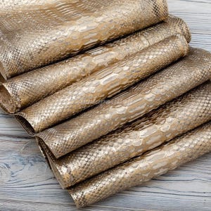 May include: A stack of folded sheets of metallic gold material featuring a raised, repeating scale pattern that mimics the texture of snakeskin. The sheets are layered on top of one another, displaying a consistent shimmering finish throughout.