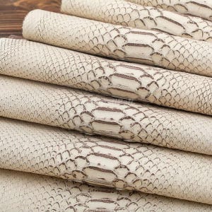 Può includere: Una pila di fogli di pelle arrotolati color bianco sporco con un motivo testurizzato a pelle di serpente. Il materiale presenta un design a scaglie ripetuto in toni crema e marrone chiaro, con una superficie goffrata uniforme sui rotoli.