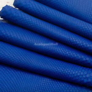 Peut inclure: Une feuille de cuir bleu roi vif présentant une texture gaufrée distincte en relief rappelant une peau de serpent sur toute sa surface. Le matériau est plié en plusieurs couches et le texte AcalapatiBali est imprimé au centre de l'image.