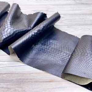 Peut inclure: Un matériau bleu marine foncé présentant un motif d'écailles répétitif et en relief sur toute sa surface. Le matériau est plié à plusieurs endroits, créant des crêtes et des courbes profondes qui soulignent la texture et le léger éclat des écailles.