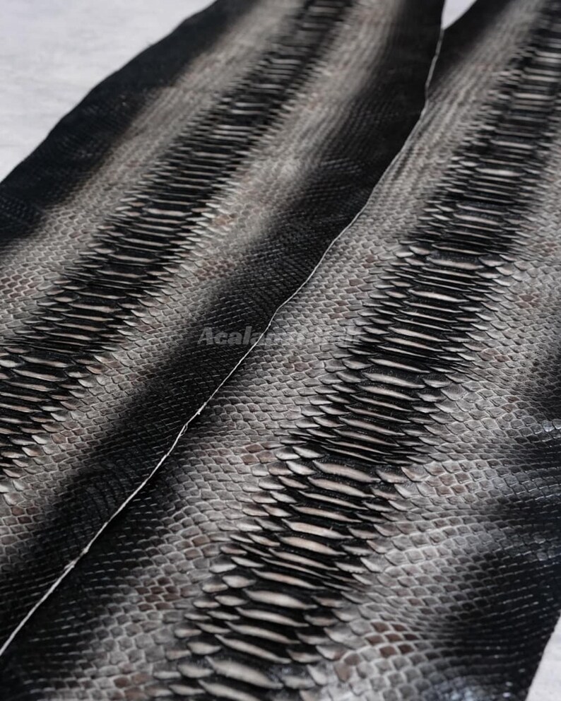 May include: Two long strips of black and grey snakeskin leather featuring a distinct scale pattern with a gradient effect. The word Acala is printed in white text across the centre of the left strip of the textured reptile hide.