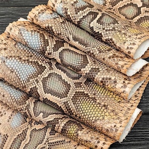 Peut inclure: Une pile de feuilles pliées en matériau texturé présentant un motif répétitif de peau de serpent marron, beige et crème. La surface affiche une texture d'écailles détaillée avec de subtils reflets bleus et jaunes irisés.