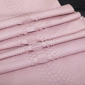 Può includere: Un foglio piegato di materiale rosa chiaro caratterizzato da una distinta trama in rilievo a pelle di serpente su tutta la superficie. Il materiale è disposto in diverse pieghe orizzontali, mettendo in mostra il motivo a scaglie e il colore tenue.