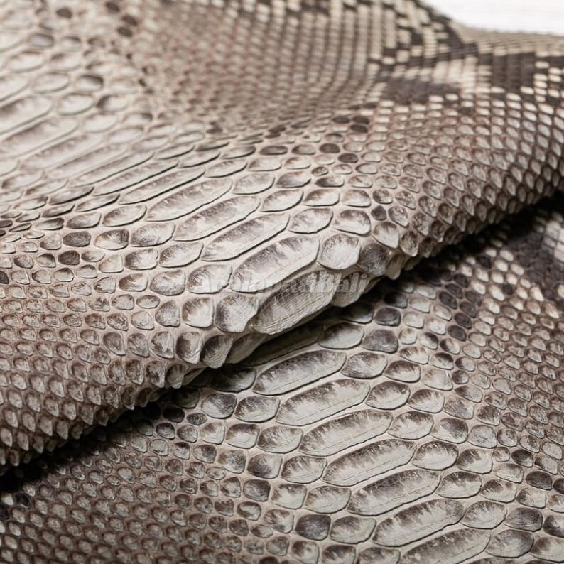 May include: A folded piece of textured snakeskin leather featuring a natural pattern of overlapping scales in shades of beige, cream, and dark brown. The surface displays a distinct organic scale structure with a matte finish and visible depth.