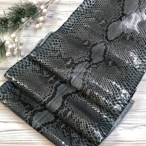 May include: A piece of grey and black snakeskin leather featuring a detailed, repeating scale pattern. The material has a glossy, reflective finish that highlights the texture of the individual scales across the entire surface of the hide.