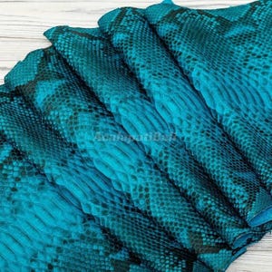 Peut inclure: Une feuille de cuir de serpent bleu sarcelle présentant un motif d'écailles en relief distinct avec des accents noirs foncés sur toute la texture. Le matériau est plié en couches, mettant en valeur les écailles complexes en forme de losange.