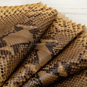 May include: A piece of tan and brown snakeskin leather featuring a distinct, repeating scale pattern with dark brown diamond-shaped markings. The material is folded in layers, showcasing the textured surface and the natural organic grain of the hide.