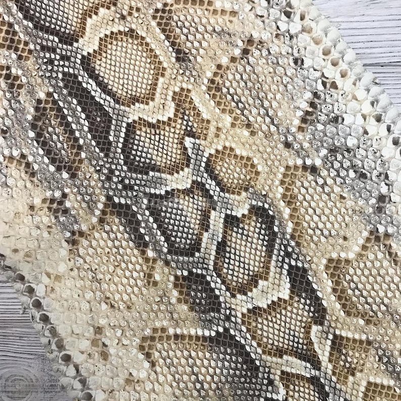 May include: A textured snakeskin material featuring a repeating geometric pattern of overlapping scales in shades of cream, beige, tan, and dark brown. The surface displays a natural, organic scale structure with a distinct central spine design.