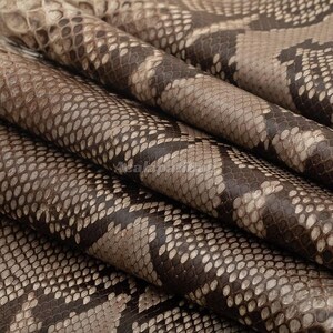 May include: A sheet of textured material featuring a repeating snakeskin pattern in shades of dark brown, tan, and cream. The surface displays a detailed, overlapping scale design with a natural, organic appearance and a matte finish throughout.