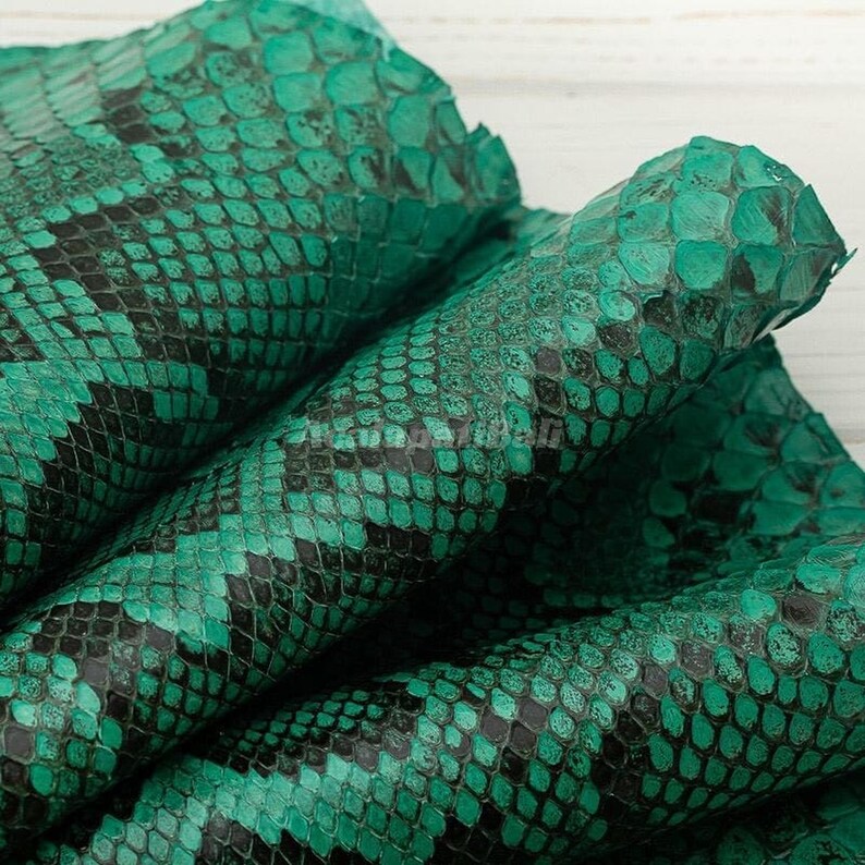 May include: A piece of genuine snakeskin leather featuring a vibrant emerald green and black scale pattern. The material has a distinct, textured surface with overlapping scales that create a repeating diamond design across the flexible hide.