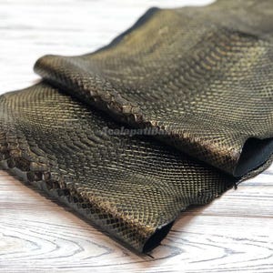 Può includere: Un pezzo di materiale scuro con una finitura oro metallizzato e un motivo testurizzato a pelle di serpente. La superficie presenta un design a scaglie ripetute che crea un aspetto dimensionale. Il materiale è ripiegato su se stesso.