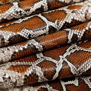 May include: A sheet of textured material featuring a repeating snakeskin pattern in shades of deep brown, tan, and off-white. The surface displays a detailed, scaled appearance with a glossy finish, arranged in overlapping, folded layers.