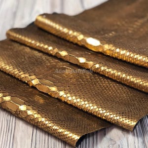 Peut inclure: Plusieurs feuilles de cuir de serpent doré métallisé avec une texture d'écailles en relief distincte. Le matériau présente une finition brillante et réfléchissante qui capte la lumière sur les couches superposées, révélant le motif naturel.