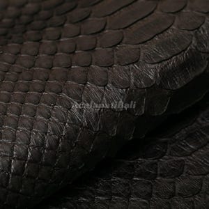 May include: A dark brown snakeskin leather material featuring a distinct, repeating scale pattern across the surface. The texture is layered and tactile, with the text "AcalapatiBali" subtly visible in the centre of the image.