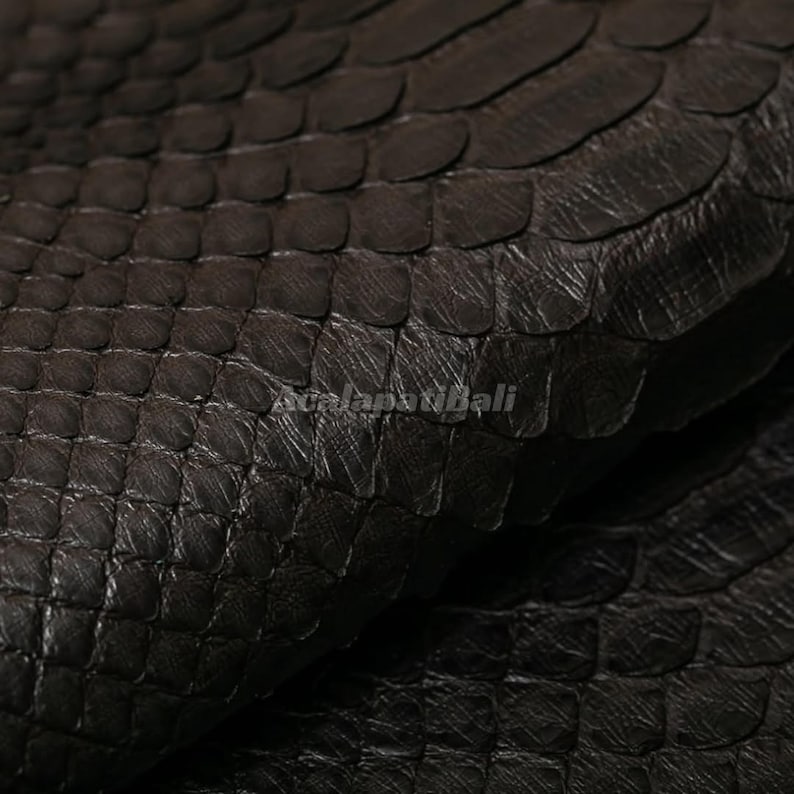 May include: A dark brown snakeskin leather material featuring a distinct, repeating scale pattern across the surface. The texture is layered and tactile, with the text "AcalapatiBali" subtly visible in the centre of the image.