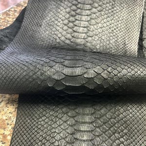 May include: A piece of black snakeskin leather featuring a distinct, repeating scale pattern. The material is folded over itself, showcasing the texture and the natural arrangement of the scales across the surface of the dark, matte-finished hide.
