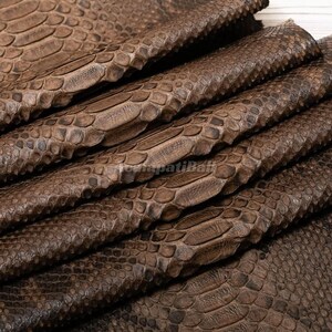 May include: A folded sheet of brown material featuring a distinct, raised scale pattern that mimics the texture of snake skin. The surface displays a gradient of dark and light brown tones, with overlapping layers creating deep, textured ridges.