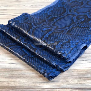 Peut inclure: Une pièce de tissu pliée présentant un motif peau de serpent bleu et noir vibrant. La texture est détaillée avec de petites écailles superposées qui créent une apparence reptilienne réaliste sur toute la surface du matériau.
