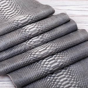 Peut inclure: Une pile de feuilles pliées en similicuir gris présentant un motif texturé en peau de serpent. La surface affiche un design à écailles répétitives avec des nuances de gris et d'argent, créant un aspect reptilien réaliste sur le tissu.