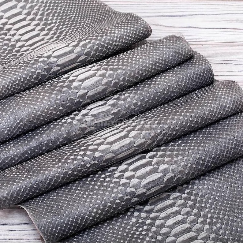 Op de afbeelding: Een stapel gevouwen vellen grijs kunstleer met een gestructureerd slangenleerpatroon. Het oppervlak toont een herhalend schubbenontwerp met verschillende tinten grijs en zilver, wat zorgt voor een realistische reptielachtige uitstraling op de stof.