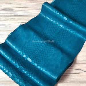 Peut inclure: Une peau de serpent en cuir de couleur bleu sarcelle vif présentant un motif d'écailles répétitif et distinct sur toute la surface. Le matériau est plié en couches, affichant une finition brillante et le texte AcalapatiBali au centre.
