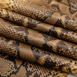 Puede incluir: Una lámina de piel de serpiente plegada con un patrón de diamantes repetitivo en tonos bronceado, marrón y gris carbón oscuro. La superficie muestra una estructura de escamas texturizada y realista con un acabado brillante y natural.