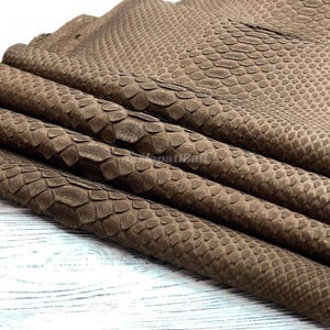 May include: A piece of brown leather featuring a textured, embossed snakeskin pattern. The material is folded in several layers, showcasing the repeating scale-like design across the surface. The color is a consistent, muted earth-tone brown.