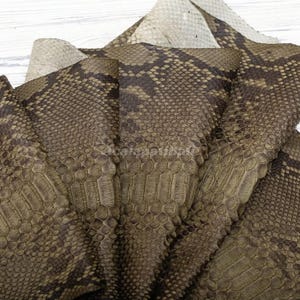 May include: A collection of overlapping sheets of textured leather featuring a dark brown and tan snakeskin pattern. The material displays a realistic scale design with a matte finish, showing the underside of the hide in a light beige colour.
