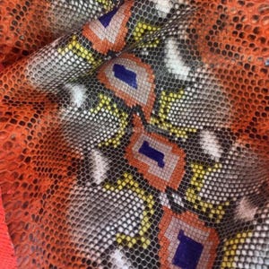 May include: A piece of textured snakeskin leather featuring a repeating geometric pattern in vibrant shades of orange, yellow, white, and deep blue. The material has a distinct scale-like surface texture with a glossy finish throughout.