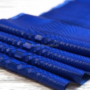 May include: A vibrant royal blue snakeskin leather sheet featuring a distinct, overlapping scale pattern across its surface. The material is folded in layers, showcasing the texture and the text AcalapatiBali printed in the center of the material.