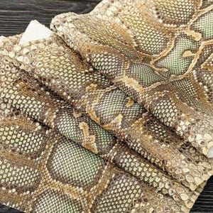 Peut inclure: Un morceau de cuir de serpent texturé présentant un motif d'écailles répétitif dans des tons de beige, de brun et de vert pâle. Le matériau est plié en couches, mettant en valeur la texture organique détaillée et le dégradé naturel de la peau.