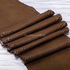 Peut inclure: Un morceau de cuir marron avec un motif texturé en peau de serpent, plié en plusieurs couches. Le matériau présente un design répétitif en forme d'écailles sur toute sa surface, avec le texte AcalapatiBali visible au centre des plis.